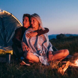 Ein Paar sitzt in der Dämmerung vor einem Zelt, ein Lagerfeuer brennt.