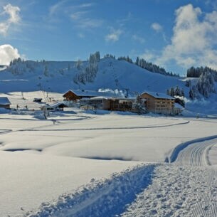 Loipen neben Neuschnee, im Hintergrund Hütten und ein schneebedeckter Berg.