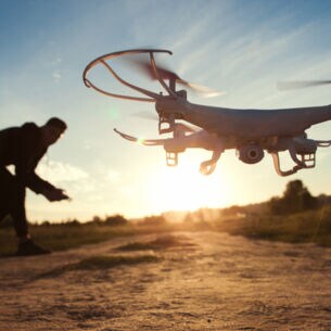Eine Drohne mit Kamera an der Unterseite fliegt im Tiefflug über ein Feld im Sonnenuntergang, im Hintergrund der Pilot als Silhouette