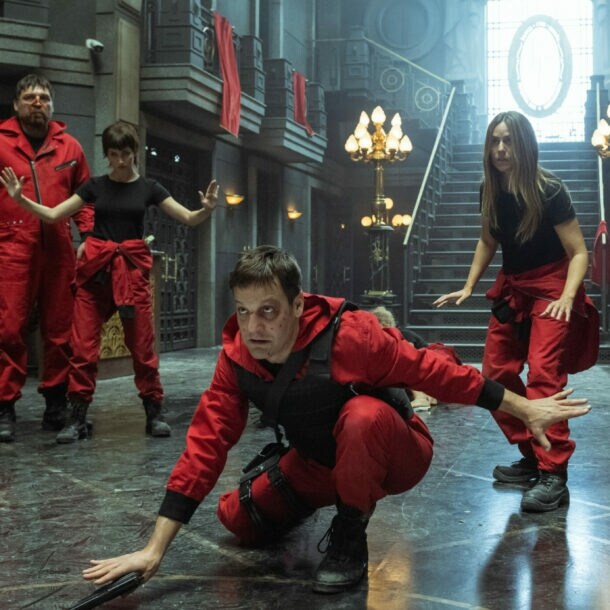 Fünf Personen in roten Anzügen in defensiver Haltung in einer prächtigen Eingangshalle aus Stein und Marmor