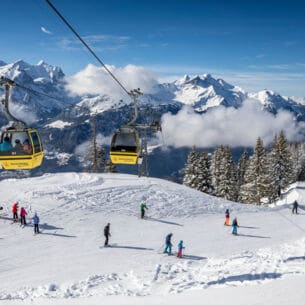 Skipiste mit Wintersportler:innen über die eine Seilbahn läuft, im Hintergrund Bergpanorama