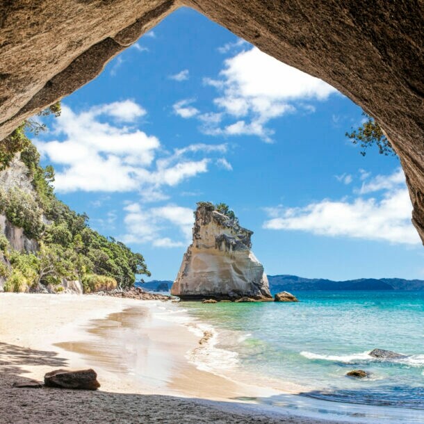 Im Vordergrund ein Felsbrocken, dahinter ein Sandstrand, umringt von Felsen und türkisfarbenem Meer