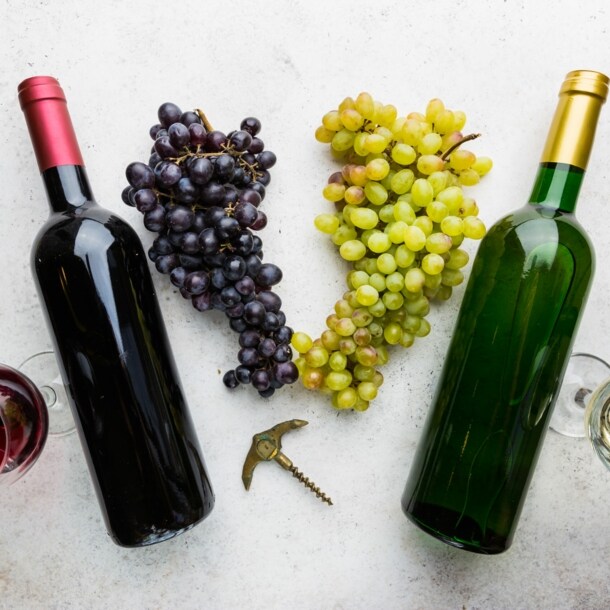 Je eine Flasche und ein Glas Rotwein und Weißwein auf einer hellen Fläche, dazwischen liegen Trauben.