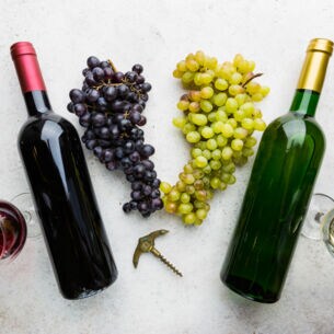 Je eine Flasche und ein Glas Rotwein und Weißwein auf einer hellen Fläche, dazwischen liegen Trauben.