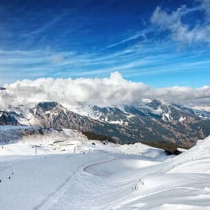 Blick von oben auf eine Piste mit Skifahrer:innen, im Hintergrund Berge