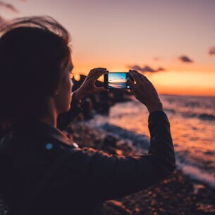 Eine junge Frau fotografiert einen Sonnenuntergang am Meer mit dem Handy