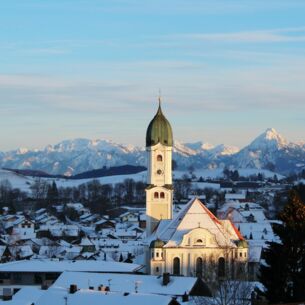 Schneebedeckter Ort mit weißer Kirche vor Alpenpanorama in der Dämmerung