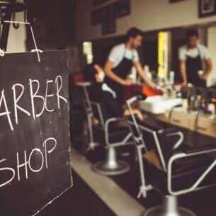 Eine Tafel auf der mit Kreide Barbershop steht, im Hintergrund ist ein Barbier mit seinem Kunden im Laden zu sehen