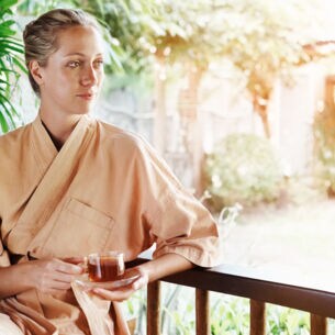 Eine junge Frau sitzt im Morgenmantel auf einer Terrasse und hält eine Tasse Tee in ihren Händen