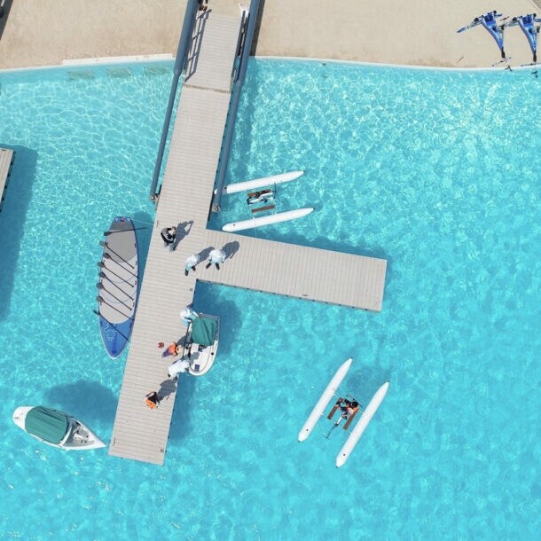 Ein großer Pool mit hellblauem, klaren Wasser sowie Booten, Katamaranen und zwei geometrischen hellgrauen Stegen in Betonoptik