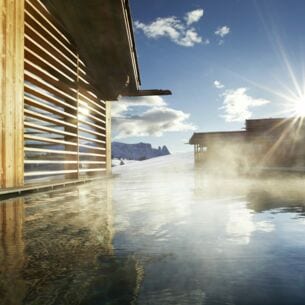 Ein Pool in der strahlenden Sonne, daneben Holzhäuser und Berge im Hintergrund