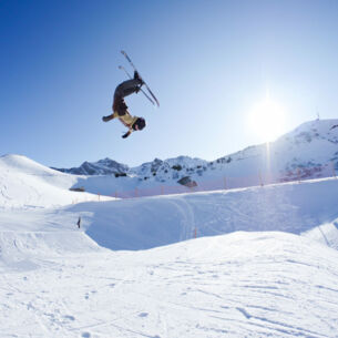Eine Person, die mit Ski einen Salto in der Luft macht, im Hintergrund ein schneebedeckter Berg