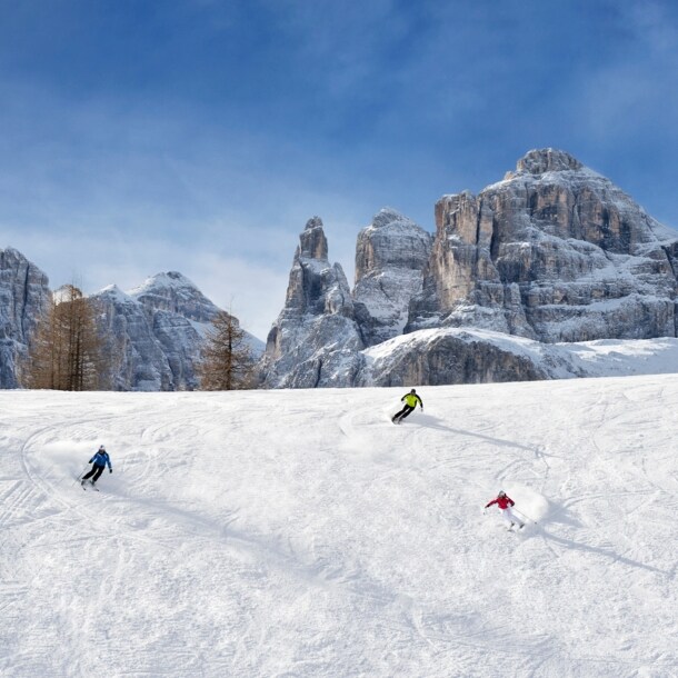 Panorama einer Skipiste mit drei Skifahrern vor markanten Felsformationen eines Bergmassivs