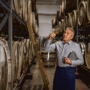 Ein Brandmeister steht in seiner Destillerie und hält ein Glas seines Whiskys prüfend gegen das Licht