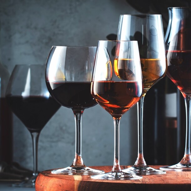 Mehrere gefüllte Weingläser stehen nebeneinander auf einem Brett, dahinter Weinflaschen vor dunklem Hintergrund.