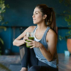 Eine junge Frau sitzt in Sportkleidung mit einem grünen Saft in ihren Händen auf einer Yogamatte in einem Raum mit Grünpflanzen.