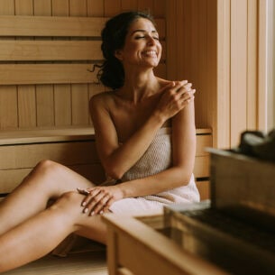Eine lachende Frau sitzt eingewickelt in ein handtuch in einer Holzsauna.
