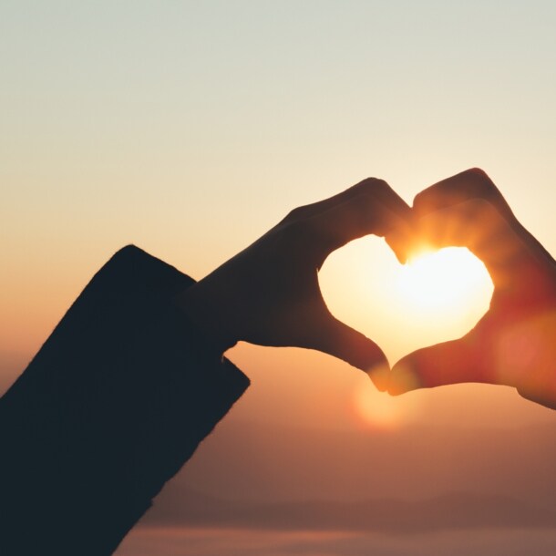 Zwei Hände formen ein Herz vor einem Sonnenuntergang