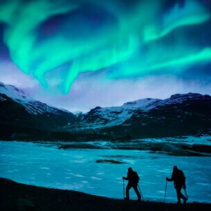 Zwei Wanderer vor einer Bergkulisse mit Polarlichtern.
