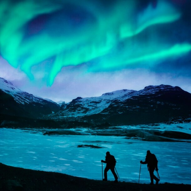Zwei Wanderer vor einer Bergkulisse mit Polarlichtern.