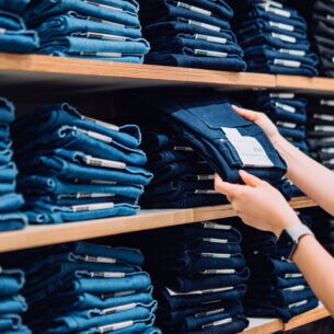 Zwei Hände greifen eine blaue Jeanshose aus einem Regal mit gestapelten Jeanshosen in einem Geschäft