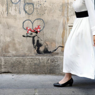 Eine Frau in langem, weißen Kleid läuft an einem Banksy-Werk vorbei: eine Ratte mit roter Minnie-Mouse-Schleife auf dem Kopf.