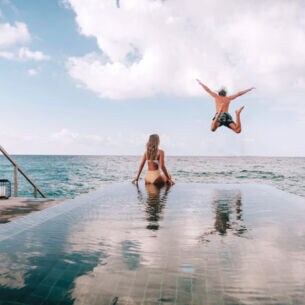 Eine Frau sitzt am Rande eines Pools am Meer, während ein Mann ins Meer springt