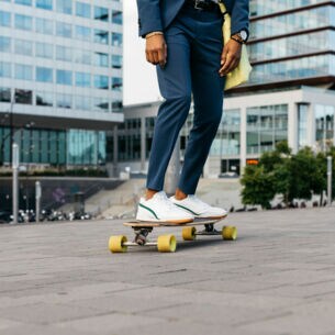 Zwei Beine auf einem Skateboard, die Person trägt eine Anzughose und weiße Sneaker