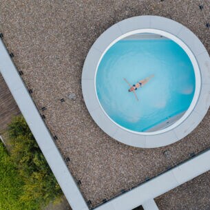 Eine Frau schwebt mit ausgestreckten Armen in einem Pool, fotografiert durch ein rundes Dachfenster aus der Vogelperspektive