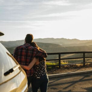 Ein Paar steht vor dem Auto und genießt die Aussicht über die Landschaft.