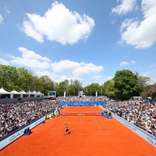 Tennisspieler spielen auf einem Sandplatz, umgeben von zahlreichen Zuschauern.