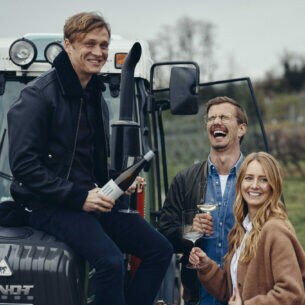 Matthias Schweighöfer sitzt lachend mit einer Weinflasche auf einem Traktor, daneben Joko Winterscheidt und Winzerin Juliane Eller auf einem Weinberg