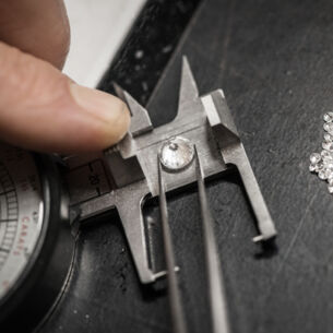 Eine Pinzette, die einen Diamanten auf die Waage legt, die von einem Finger gehalten wird, daneben liegen kleine Diamanten