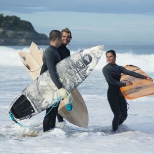Drei Menschen stehen in Neoprenkleidung und Surfbrettern im Wasser.