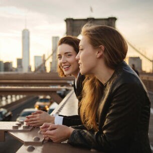 Zwei junge Mädchen stehen lächelnd auf der Brooklyn Bridge vor der Skyline Manhattans