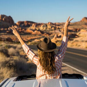 Eine junge Frau ragt mit dem Oberkörper aus dem Schiebedach eines Autos auf einer Straße in einer Wüstenlandschaft und winkt spontan mit einem Hut in die Luft