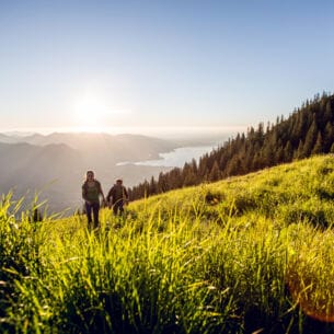 Ein Paar wandert einen grünen Hügel hinauf, im Hintergrund scheint die Sonne und ist ein See zu erkennen.