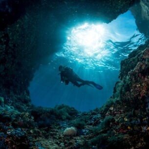 Taucher unter Wasser am Eingang einer Höhle mit Sonnenstrahlen im Hintergrund