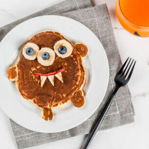 Frühstücksteller mit einem Pancake in Form eines kleinen Monsters, dessen Augen aus Bananen und Blaubeeren bestehen
