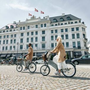 Zwei schick gekleidete Personen auf Fahrrädern vor dem neoklassizistischen Gebäude des Hotel d´Angleterre in Kopenhagen