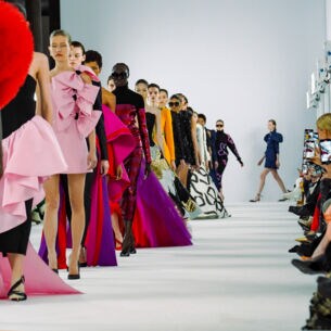 Eine Reihe weiblicher Models in bunten Abendkleidern vor Publikum auf einem Laufsteg