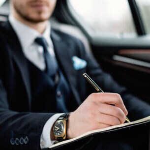 Ein Mann im Anzug sitzt in einem Auto und notiert etwas mit einem Kugelschreiber in einem Notizbuch