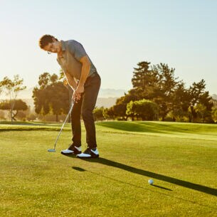 Ein Mann steht auf einem Golfplatzt und locht gerade den Golfball ein.