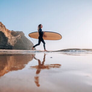 Eine Frau im Neoprenanzug trägt ihr Surfbrett unterm Arm ins Meer