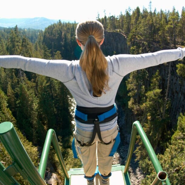 Eine Frau steht mit ausgebreiteten Armen gesichert an einem Bungee-Jumping-Gerüst und ist kurz davor runterzuspringen