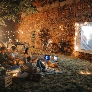 Eine Personengruppe sieht mit einem Mini-Beamer einen Film an einer Mauer im Garten.