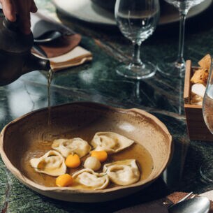 An einem stilvoll gedeckten Restauranttisch läuft Brühe aus einer Kanne in einen Teller mit gefüllter Pasta