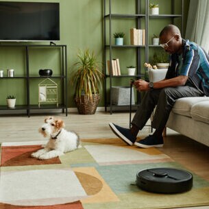 Ein Mann sitzt im Wohnzimmer auf dem Sofa und schaut auf sein Handy in der Hand. Vor ihm liegt ein Hund auf dem Teppich, daneben arbeitet ein Saugroboter.
