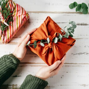 Ein Geschenk, verpackt in einem orangefarbenen Tuch und geschmückt mit einem grünen Zweig, wird von zwei Händen gehalten.