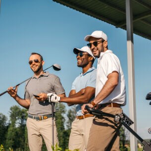 Drei Männer in Poloshirts, mit Sonnenbrillen und Golfschlägern stehen auf einer überdachten Terrasse und lachen